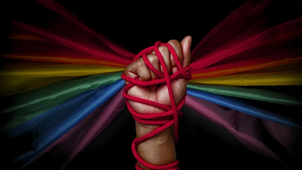 Fist tied in red bondage rope in front of a rainbow flag